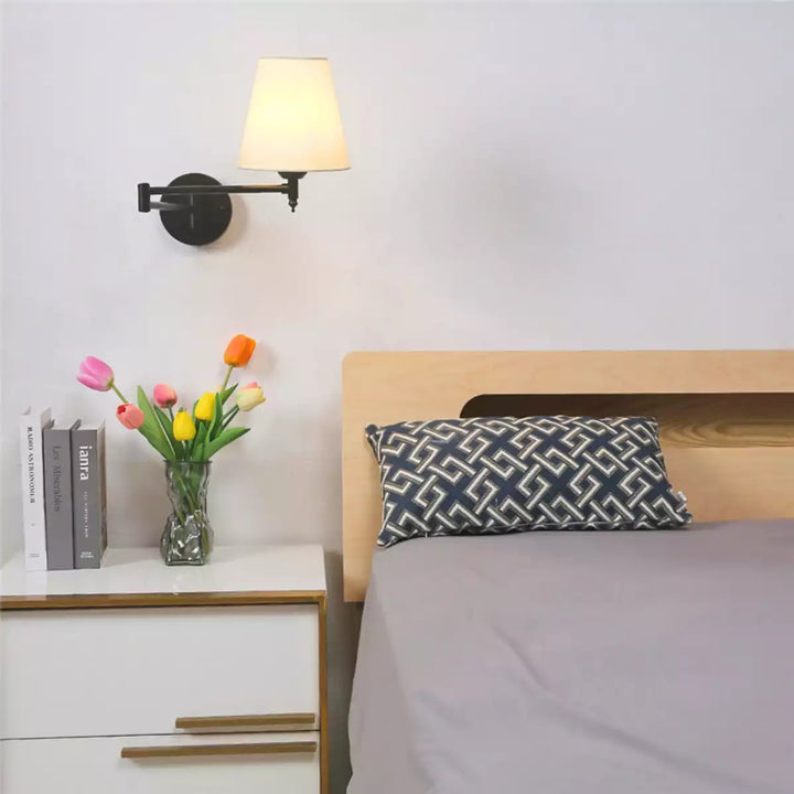 Luminária de parede articulada preta em um quarto ao lado de uma cama com travesseiro decorativo