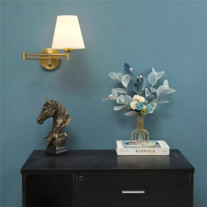 Luminária de parede articulada dourada instalada em parede azul ao lado de uma cômoda preta com decoração
