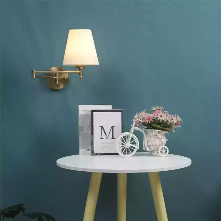 Luminária de parede articulada dourada em parede azul sobre uma mesa lateral branca com objetos de decoração