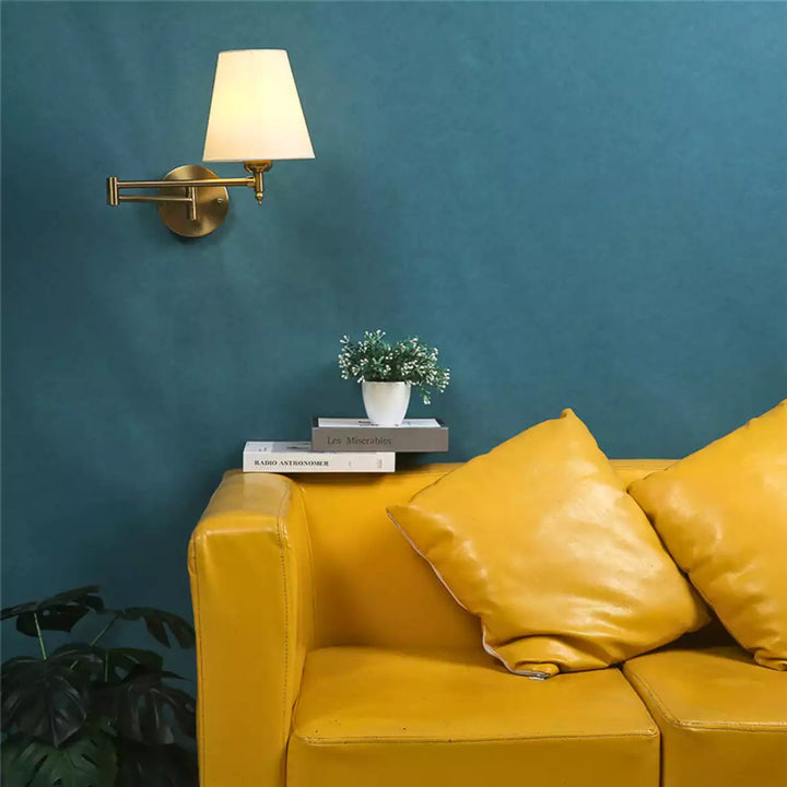 Luminária de parede articulada dourada em parede azul sobre um sofá amarelo com almofadas